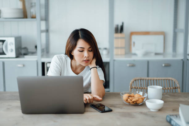istock Young Asian woman working from home using laptop computer while reading text messages on smartphone in the kitchen at home. Remote working, freelancer, small business concept 1366367226