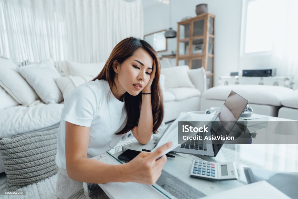 Young Asian woman handling home finances with laptop, looking worried while going through financial bills. Financial plan, tax, spending and budgets, financial problems concept Worried Stock Photo