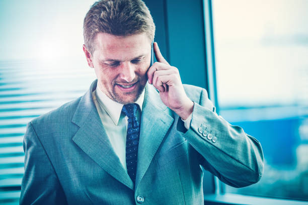 Happy businessman talking on the smart phone standing near big window. A view through the glass wall with white stripes.