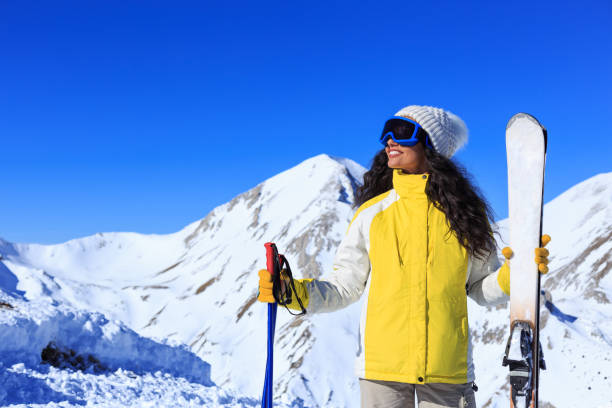 노란 재킷을 입은 여성은 미소를 짓고 겨울 휴가를 즐기며 밝은 푸른 하늘이 있는 산 배경에 스키 장비를 들고 있습니다. 스톡 사진