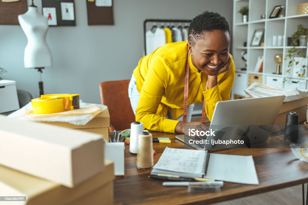 A professional young woman who leans on a desk and works on a laptop African professional young woman works on a laptop in her large home office. African-American Ethnicity Stock Photo