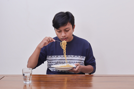 Portrait of hungry Asian man in casual sweater eating instant noodles using fork on the table