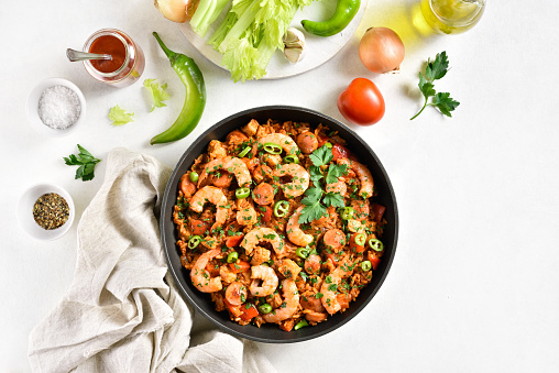 Creole jambalaya with chicken, smoked sausages and vegetables in frying pan on white stone background. Top view, flat lay