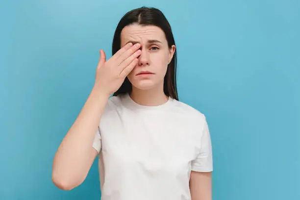 Photo of Young girl suffering from eyes pain and feeling something in eye, posing on blue wall. Cause of pain include contact lens problem, conjunctivitis, foreign object, dry eye syndrome or allergy