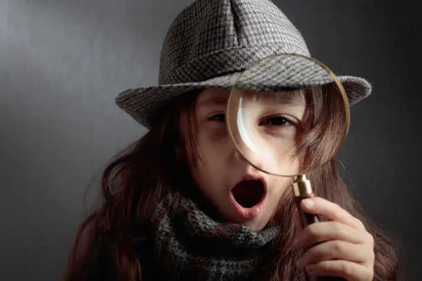 The surprised little detective with a magnifying glass. A little girl in a hat and coat is playing detective. Copy space.