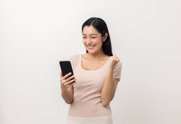 istock Excited Beautiful young asian woman using smartphone mobile standing on isolated white background. Playing game on smartphone winning victory moment. Very happy enjoy and fun relax time 1366400448
