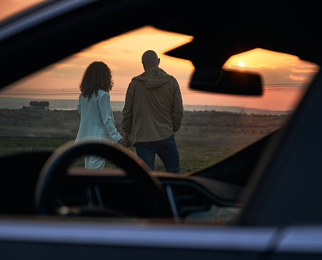 View through open side and front car window of couple in love. Back view of romantic pair looking at evening sky in orange and red colors while sunset out of city.