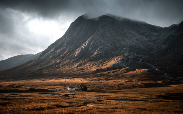 Dark clouds gathering over the peak of Buachaille Etive Mor Mountain in Glencoe, Scottish Highlands. The clouds give the mountain a moody, dramatic look. The small White House lies in the shadow of the mountain. High Quality Photo.