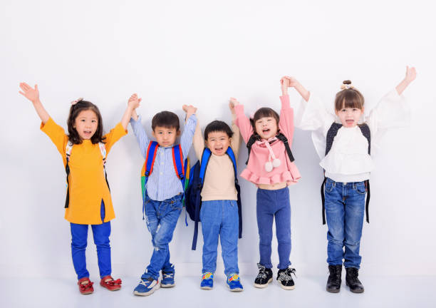 A multi-ethnic group of preschool kids is indoors in their classroom. They are sitting on the floor and raising their hand to answer a question posed by their teacher while reading a story. They are all smiling.