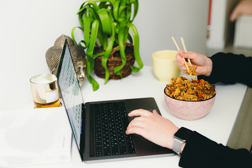 Young woman sitting at the table working and studying by laptop at hew white-designed apartment, and having delivered Asian dinner with traditional Thai fried rice with shrimps
