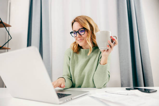 A happy entrepreneur working online on her laptop from home office and drinking coffee.