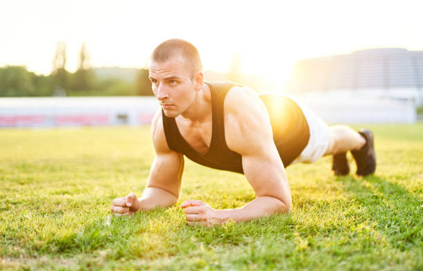 muscular concentrated young man holding plank position outdoors in the grass in sunset - morning warm up - motivation and healthy lifestyle concept with an athletic 20s sportsman - playing field effort outdoors human age imagens e fotografias de stock