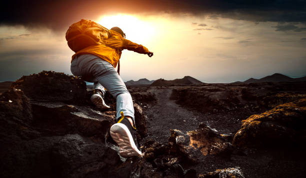 hombre exitoso caminando montañas al atardecer - excursionista con mochila apuntando al cielo con el dedo - determination fotografías e imágenes de stock