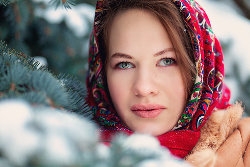 Beautiful Slavic woman, in winter, in a headscarf on her head, looks out into the distance, against the background of coniferous trees. Russian winter, Big red lips. Snow falls