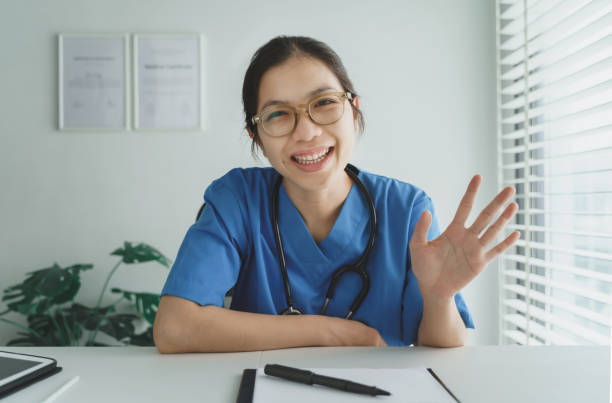 smiling asian female doctor looking at webcam making conference video call - doctor patient greeting talking imagens e fotografias de stock