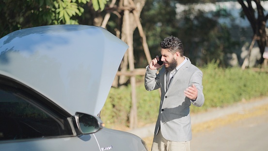 Irritable businessman in suit standing speak on phone for roadside assistance near the car opened the hood. A young man with broken car on the road need help to repair. Stress problem emergency insurance auto.
