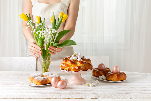 Happy Easter! Woman preparing table with Easter pastry, eggs, candies and spring flowers for holiday. Light background. Copy space.