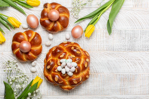 Easter background with italian style braid bread and pink metallic  colored eggs, candies. Homemade pastry. Yellow Tulips. White wooden background. Top view, copy space.