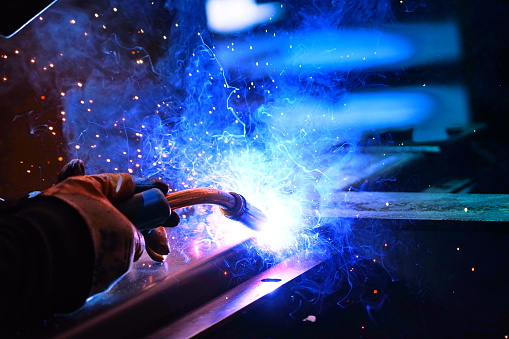 A welder welds metal into his workshop. Blue welding sparks. Gas combustion and blue smoke. Small welding workshop. Welding juncture of metal construction