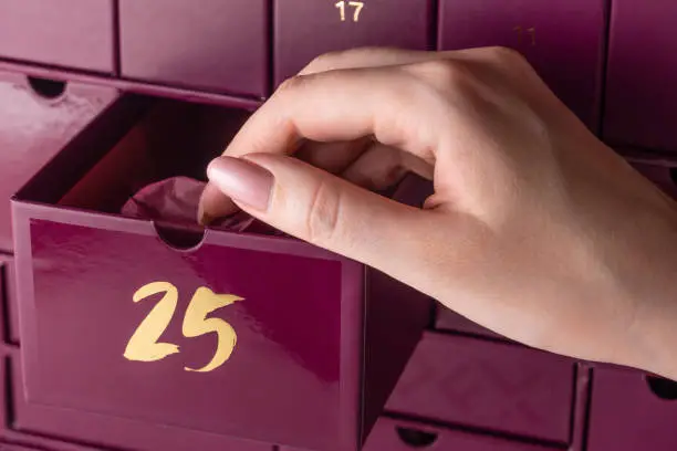 A woman's hand with a manicure opens the cell number 25 in the advent calendar. The girl takes out a gift in wrapping paper from the advent calendar.