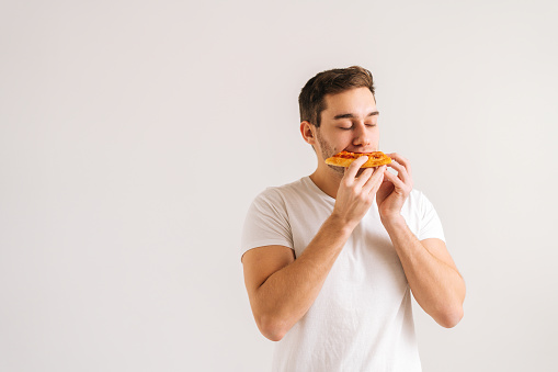 Portrait of happy young man with enjoying eating delicious slice of pizza, with closed eyes from pleasure on white isolated background. Studio shot of hungry handsome male student eating tasty food.
