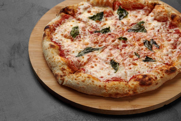 Uncut Italian pizza served with fresh basil leaf on Wooden board on dark texture background. Hot pizza with tomatoes and pesto sauce