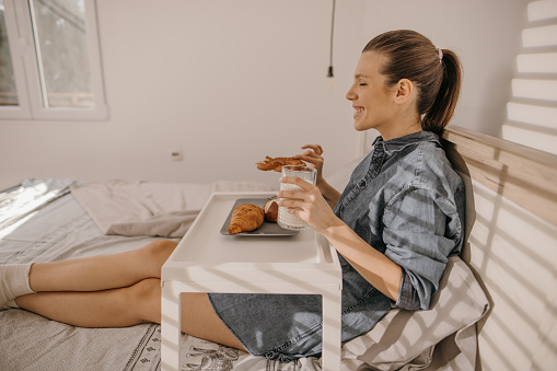 Attractive woman having pretzel and milk for breakfast, she is sitting in bedroom on a bed.