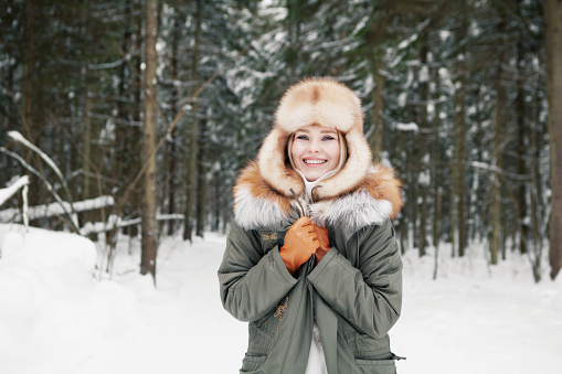 Smiling young woman in khaki parka, ear flaps ushanka hat and leather gloves at snowy coniferous forest background. Happy blonde model with beautiful smile and blue eyes in winter clothes