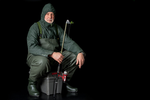 A fisherman in waders and raincoat sits on his fishing box against a black background and holds a fishing rod in his hand.