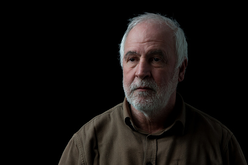 Portrait of an older gray-haired man with a beard in front of a black background.