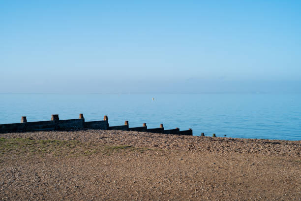 Wooden groyne water breakers on a pebble beach in Whitstable on a bright winter day with blue sky and blue sea. Isle of Sheppey can fairly be seen on the horizon.
