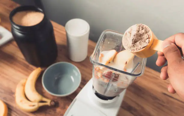 Photo of Hand holding a protein scoop over blender with smoothie.