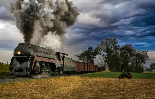 An Antique Restored Steam Freight Train Approaching Head on Blowing Smoke and Steam With an Antique Farm Tractor Watching