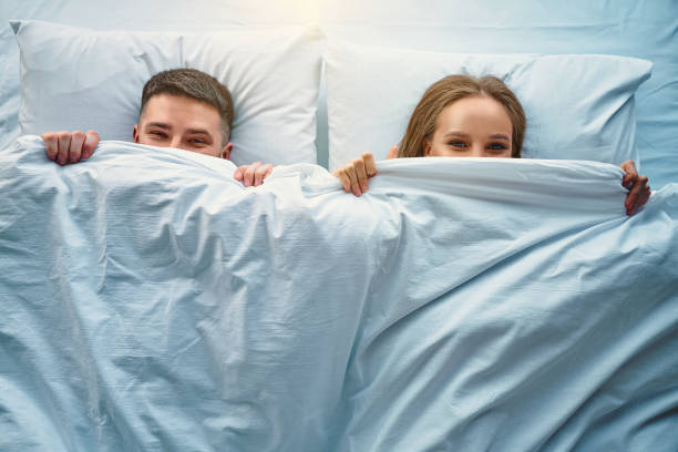陽気なカップルはベッドに横たわり、毛布の半分の顔で覆われて笑っています。喜び、狡猾さと陽気の遊び心の感情。トップビュー。 - sexuality ストックフォトと画像
