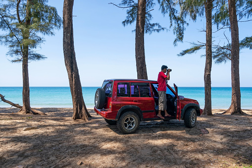 Young Asian male traveler and photographer standing on the car 4x4 car and Taking photo sea beautiful beach in Phuket Thailand Travel photography concept