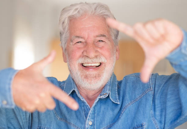 istock Happy adult mature senior bearded man looking at camera gesturing frame size with hands. Smiling laughing carefree attractive grandfather inside home with denim shirt relaxed enjoying retirement 1366504841