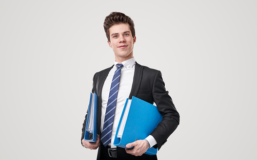 Positive male entrepreneur in formal suit standing with blue folders on gray background and looking at camera