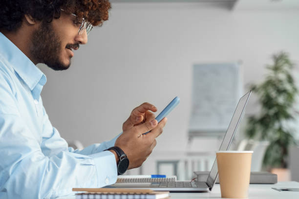 istock Indian man using mobile phone app sitting front of laptop on desk side view shot 1366517619