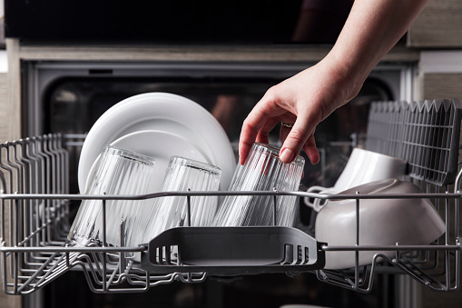 Close-up of female hand loading dished to, empty out or unloading from open automatic stainless built-in dishwasher machine with clean utensils inside in modern home kitchen. Household domestic life