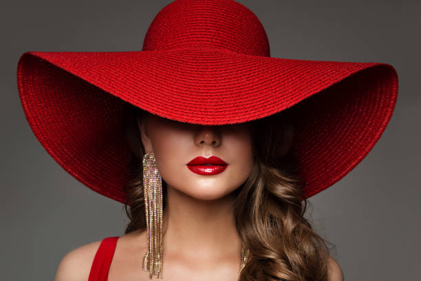 modna kobieta w kapeluszu z makijażem na czerwone usta i złotym kolczykiem. twarz modelki piękności ukryta przez szeroki szeroki kapelusz z rondem. elegancka lady portret z bliska nad szarym - big lips zdjęcia i obrazy z banku zdjęć