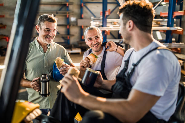 Happy manual workers and their manager talking on lunch break in a factory. Group of workers communicating while having a lunch break in industrial building. construction lunch break stock pictures, royalty-free photos & images
