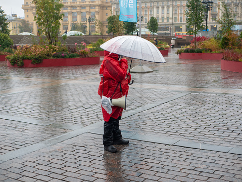 Moscow, Russia - September 21, 2021: Woman in a bright red raincoat under an umbrella on Manezhnaya Square offers to buy a bus tour of Moscow on an autumn rainy day.