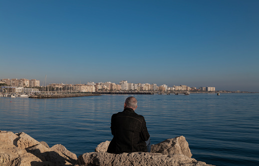 Rear view of adult man sitting on rock with lighthouse and city in background. Almeria, Spain