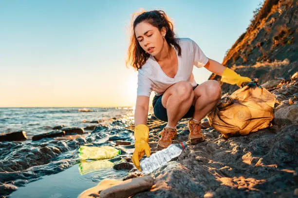 Photo of A young woman volunteer squats and picks up garbage on the ocean shore. Cleaning of the coastal zone. Sunset in the background