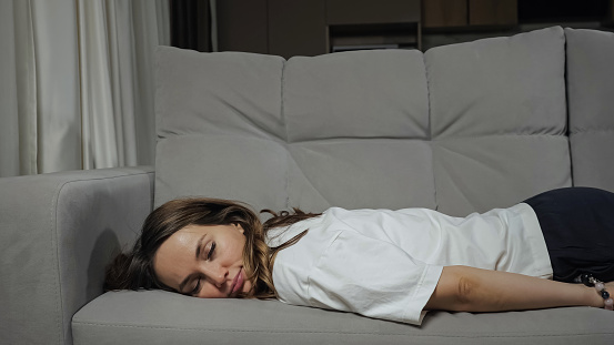 Young tired brunette woman in white t-shirt comes late from work falling on belly to comfortable grey sofa in living room at home, close view.