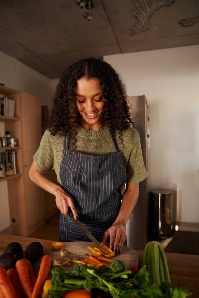 orientamento verticale di una giovane donna multiculturale che taglia le verdure su un tagliere di legno in cucina. fresco e sano - ewan foto e immagini stock