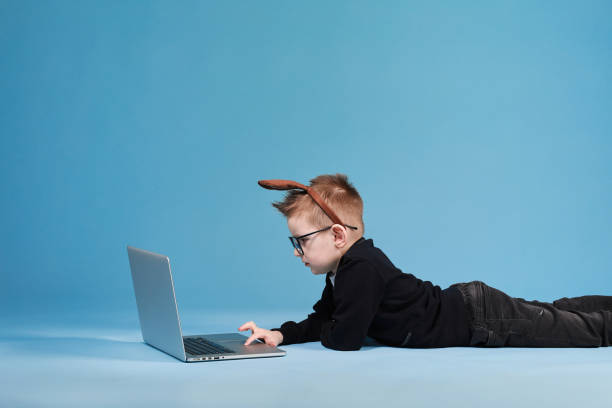 istock Distance learning online education. Schoolboy in rabbit bunny ears on head studying at home with laptop and doing school homework on blue background 1366564166