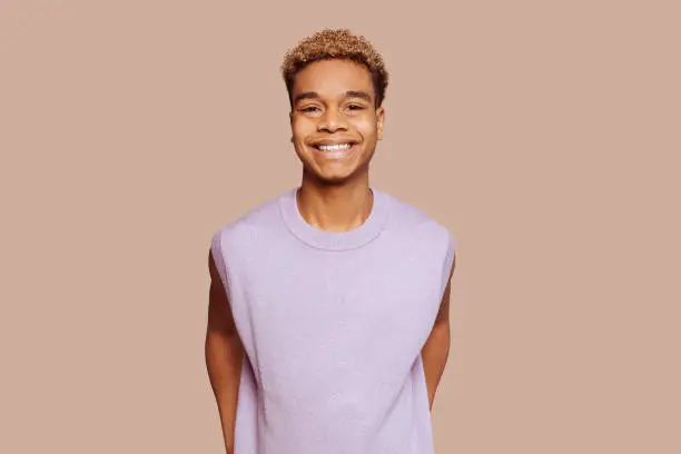 Glad young afro latin american black handsome man feels great, wears in purple top, broadly smiles and looks in camera stands over beige background.