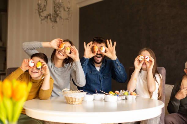 la famiglia di quattro membri è seduta al tavolo da pranzo sorridendo e tenendo le uova di pasqua vicino agli occhi. - easter easter egg paintbrush holding foto e immagini stock