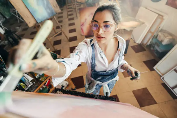 Artist painting while wearing tinted eyeglasses. Imaginative young painter making a new artwork for her project. Creative female artist standing in front of a canvas in her atelier.